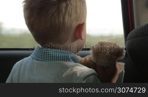 Slow motion close-up shot of a a little boy traveling by car with favourite toy bear. He hugging soft friend and they enjoying view through the window
