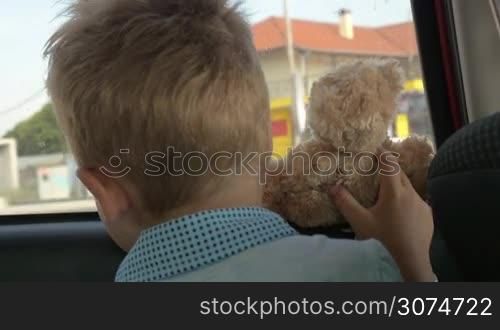 Slow motion close-up shot of a little boy with toy bear looking out the window when traveling by car, back view. He and his teddy bear are very curious about outside