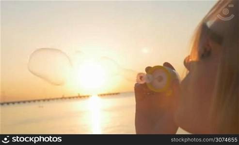 Slow motion close-up shot of a young smiling blonde blowing bubbles on the beach. Scene with sea in warm light of sunset