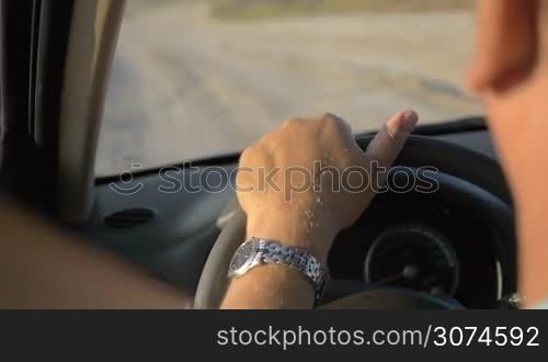 Slow motion close-up shot of man driving a car in the countryside in summer evening. View to the road and hands on the steering wheel