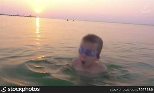 Slow motion of a boy in goggles bathing in the sea at sunset. He taking breath and diving for a moment, then he shouting with hands up as a winner