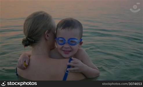 Slow motion of a boy in goggles embracing dear mother while they bathing in the sea. Mom kissing son and they looking at water. Happy family vacation