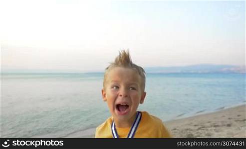 Slow motion of a boy screaming from happiness to be the winner. He wearing a medal and raising a trophy. Shot on the beach against sea and sky background