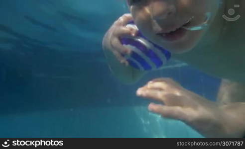 Slow motion of a boy with ball coming up from the water in swimming pool. Having fun on summer resort