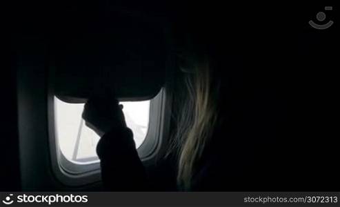 Slow motion of a female passenger of airplane opening the blind and looking out the illuminator where wing can be seen