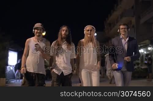 Slow motion of a group of friends with tree men and a woman walking along the night street and listening to music in headphones