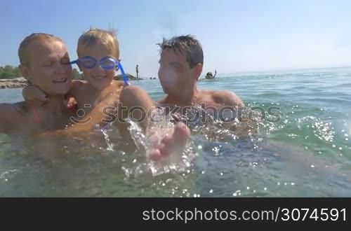 Slow motion of a little boy having enjoyable time in sea with parents. Mother and father holding him while playful child splashing water to the camera. Fun on summer holidays