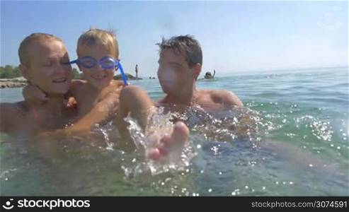 Slow motion of a little boy having enjoyable time in sea with parents. Mother and father holding him while playful child splashing water to the camera. Fun on summer holidays