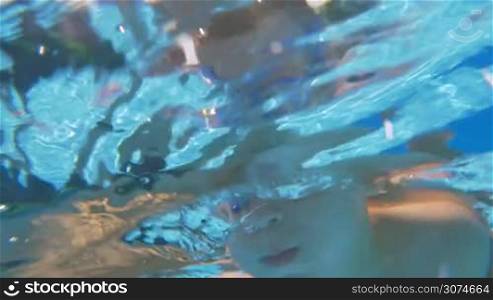 Slow motion of a little boy in goggles diving and swimming underwater in outdoor pool , then he coming up with following view of bright sunshine in clear blue sky