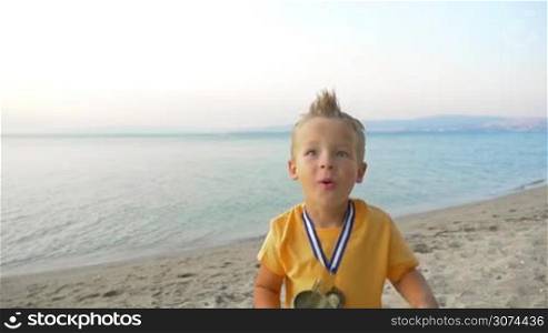 Slow motion of a little boy jumping and getting excited with the victory. He holding a cup and wearing a medal. Winner emotions, shot on the background of sea and sky