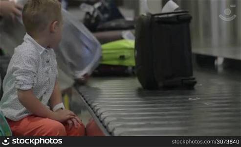 Slow motion of a little boy sitting by the baggage conveyor belt at the airport and looking at people taking luggage