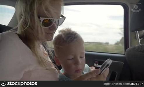 Slow motion of a little boy using mothers smart phone while they traveling on the backseat of a car. Entertainment during the ride