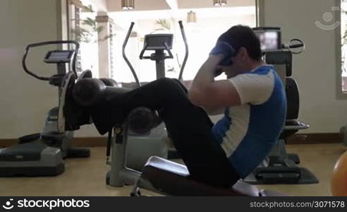Slow motion of a man exercising on modern equipped fitness center. He doing sit-ups on abdominal bench and then stopping for a break