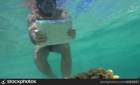 Slow motion of a man in snorkel underwater using pad in water-proof case to take pictures or shoot video of beautiful coral reef