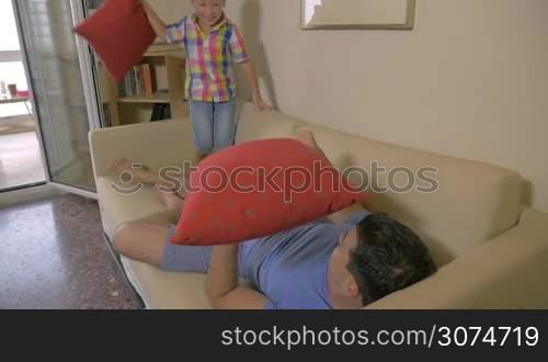 Slow motion of a son and father fighting with pillows on the sofa in the living room. Playful and naughty child having fun with beloved dad