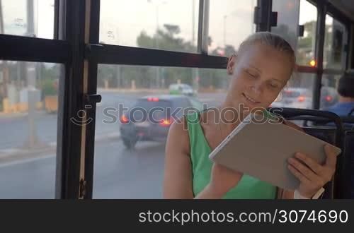 Slow motion of a young blonde woman working with tablet computer during bus ride in the city. She sitting by the window with road traffic view