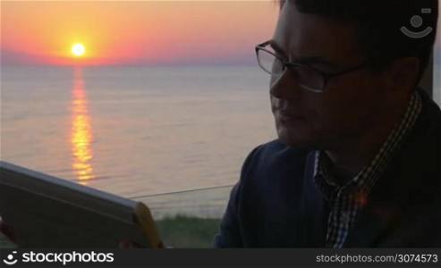 Slow motion of a young businessman using tablet PC and smoking while sitting at the balcony with view to the sea and sunset