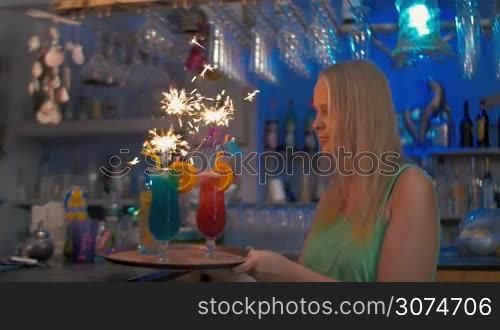 Slow motion of a young happy woman carrying the tray with cocktails in the bar. Sparklers burning in the glasses creating festive atmosphere