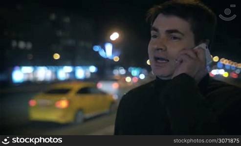 Slow motion of a young man having a phone talk in the city street at night. Defocused road with traffic and urban lights in background