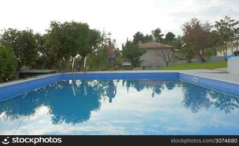 Slow motion of a young man running and jumping into the pool. Big splashes in the water. Fun and cooling on hot summer day. Vacation at villa