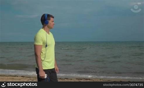 Slow motion of a young man standing on the beach and listening to music in headphones with smartphone. Nature scene with sea and sky