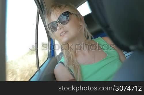 Slow motion of a young smiling woman in sunglasses traveling on backseat of a car. She looking out the window, wind blowing to the face and waving blond hair