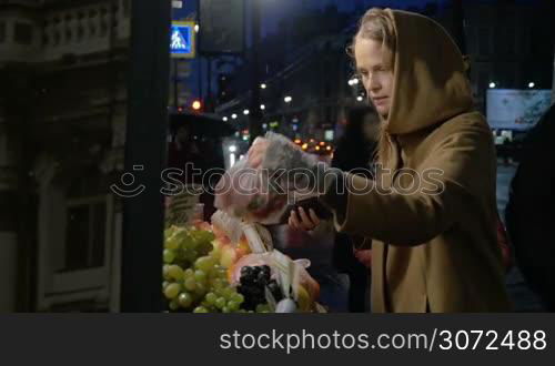 Slow motion of a young woman buying fresh strawberries and pomegranates in outdoor fruit market in late rainy evening