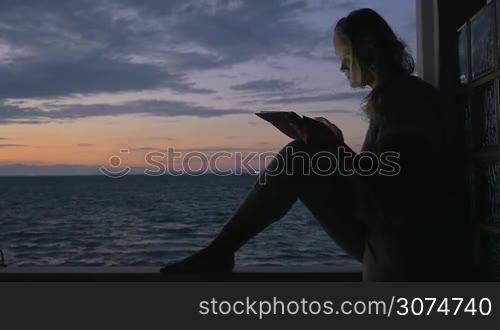 Slow motion of a young woman using tablet computer sitting on the wooden balcony rail of a seaside house. Light breeze waving her hair, twilight sky and sea in background