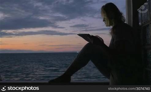 Slow motion of a young woman using tablet computer sitting on the wooden balcony rail of a seaside house. Light breeze waving her hair, twilight sky and sea in background