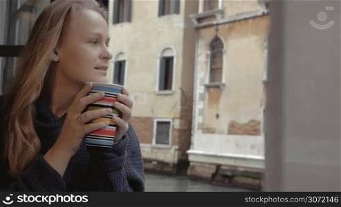 Slow motion of a young woman with tea sitting on the windowsill at home in Venice. She relaxing with hot beverage looking at scene outside