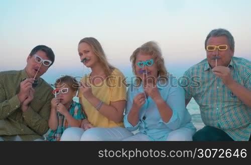 Slow motion of big hipster style family sitting on the beach by the sea. Happy parents, child and grandparents holding paper glasses and moustache