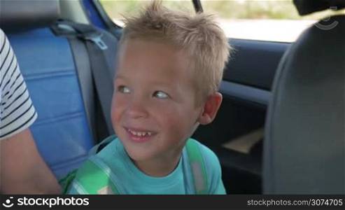 Slow motion of cute blond little boy sitting on backseat of a moving car. Happy child with rucksack looking forward new traveling experience