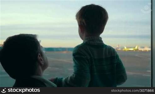 Slow motion of father and son looking out the window with view on airport area. Truck passing by, planes can be seen in the distance