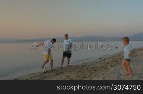 Slow motion of father, son and grandfather playing football on the beach in the evening. Family team in blue and white striped t-shirts