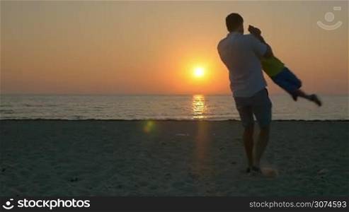 Slow motion of father spinning son on the beach. Child and dad having fun time on the shore during beautiful sunset over sea