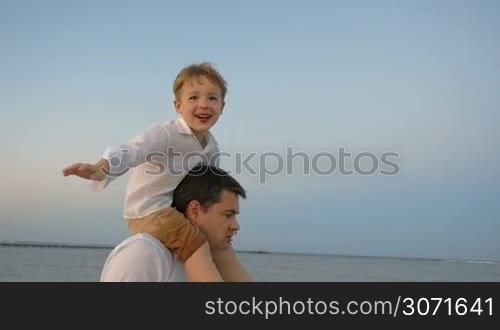 Slow motion of happy little boy sitting on fathers shoulders and imitating plane while they walking by the sea