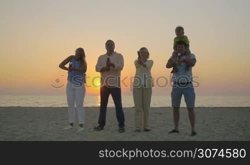 Slow motion of happy parents and grandparents making confetti firework with crackers on the coast at sunset. Little boy sitting on fathers shoulders. Family celebration