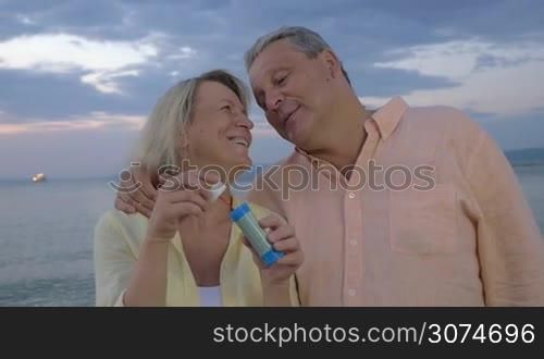 Slow motion of happy senior man and woman blowing soap bubbles on the shore. Cloudy evening sky and sea in background