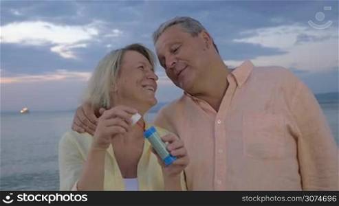 Slow motion of happy senior man and woman blowing soap bubbles on the shore. Cloudy evening sky and sea in background