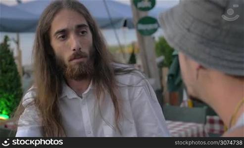 Slow motion of long-haired man with beard talking to his friend in outdoor cafe, he holding lit cigarette in hand