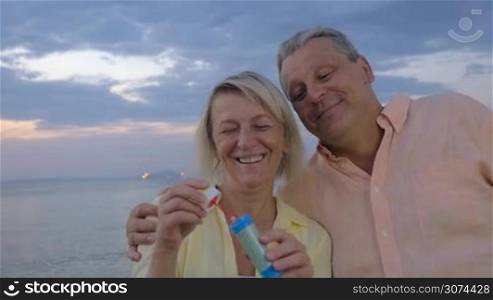 Slow motion of mature couple spending evening at the seaside. Woman blowing bubbles and they watching them flying up in the sky