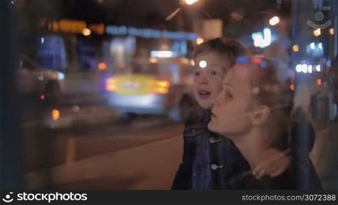 Slow motion of mother and son at bus stop by the road with intense traffic at night. Boy looking into distance and pointing at something. View through the glass