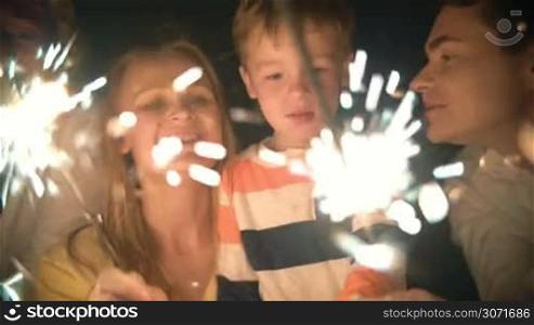 Slow motion of parents with sparklers and son watching them at night. Father and mother kissing child and he kissing them. Family celebration