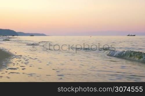 Slow motion of sea waves washing the shore at sunset, boats in the distance. Scenic seascape with sky in warm evening colors
