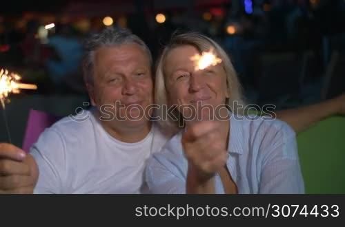 Slow motion of senior couple with Bengal lights sitting outdoor at night. They waving with sparklers and smiling