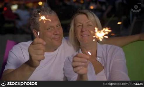 Slow motion of smiling senior man and woman waving with sparklers sitting outdoor at night