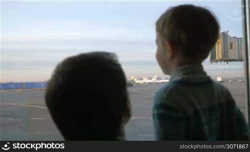Slow motion of son and father by the airport window. The looking at takeoff strip with trucks and planes