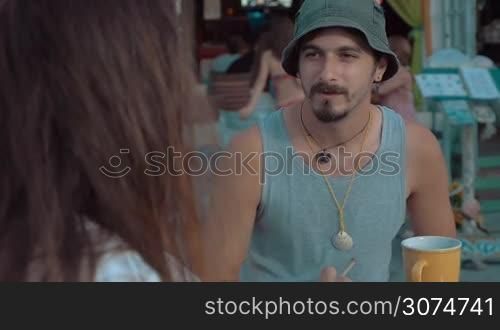 Slow motion of two friends sitting in outdoor cafe. Young man with beard wearing bucket hat listening to his smoking long-haired old chap