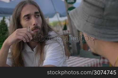 Slow motion of two male friends sitting in outdoor cafe. They smoking and having a talk. One man is long-haired with beard, another is wearing a bucket hat