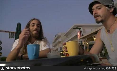 Slow motion of two male friends toasting with mugs and having drinks in street cafe on resort in the evening